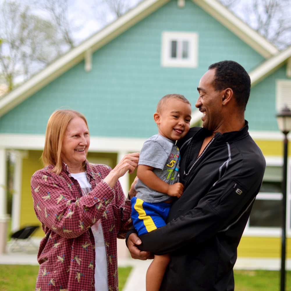 An older African American man is smiling holding a young African American boy in his arms. There is an older caucasian woman standing beside them smiling. Behind them is a blue and yellow home.