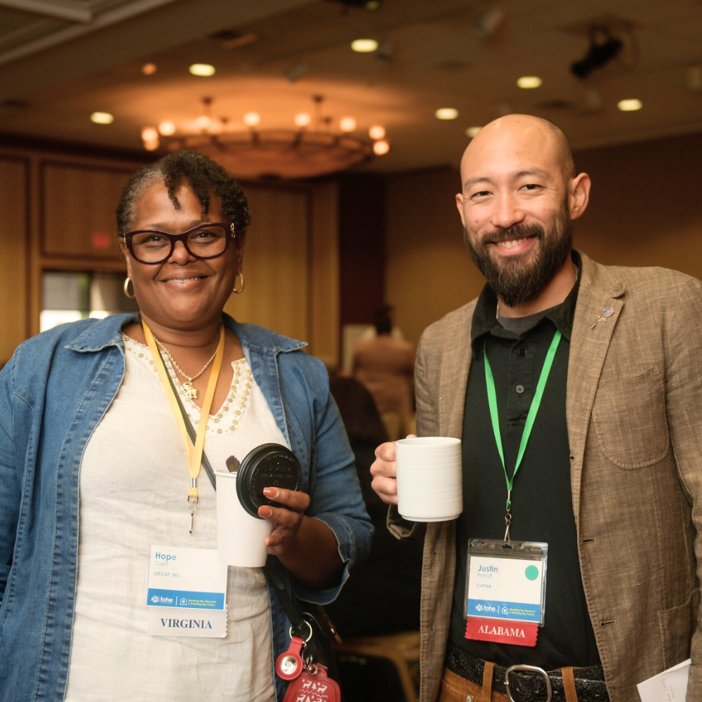 A middle aged African American woman and asian man are standing together smiling holding coffee cups in a ballroom. They have Fahe member lanyards around their necks.