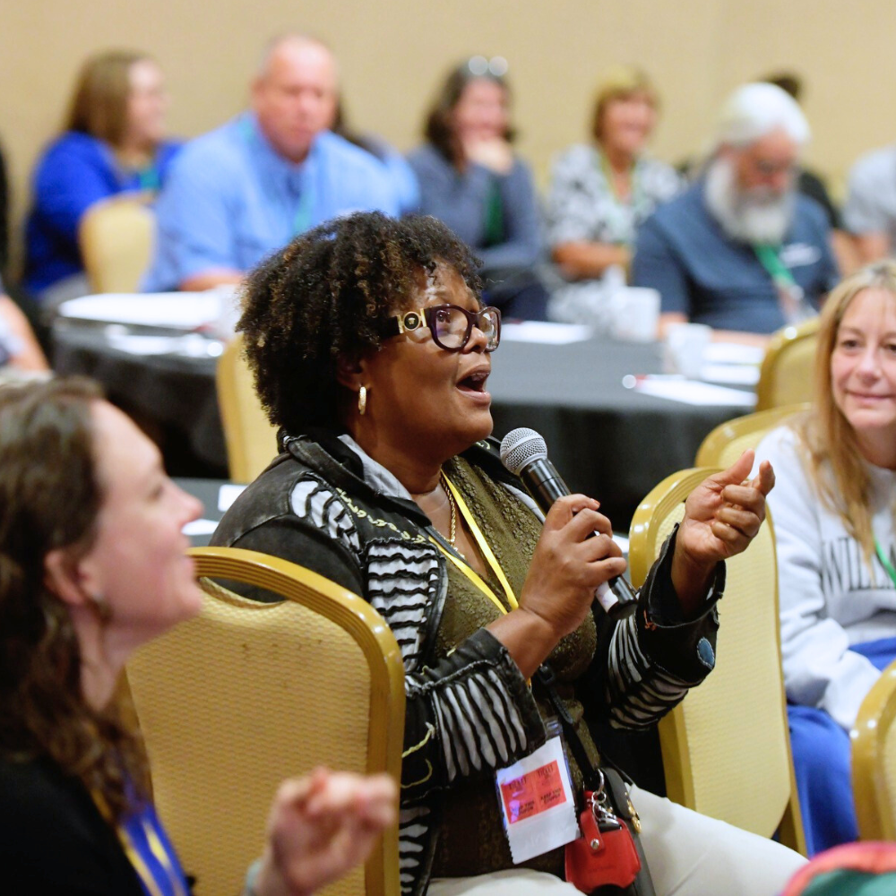 An African American woman is sitting speaking into a microphone. There are people seated around her at tables listening.