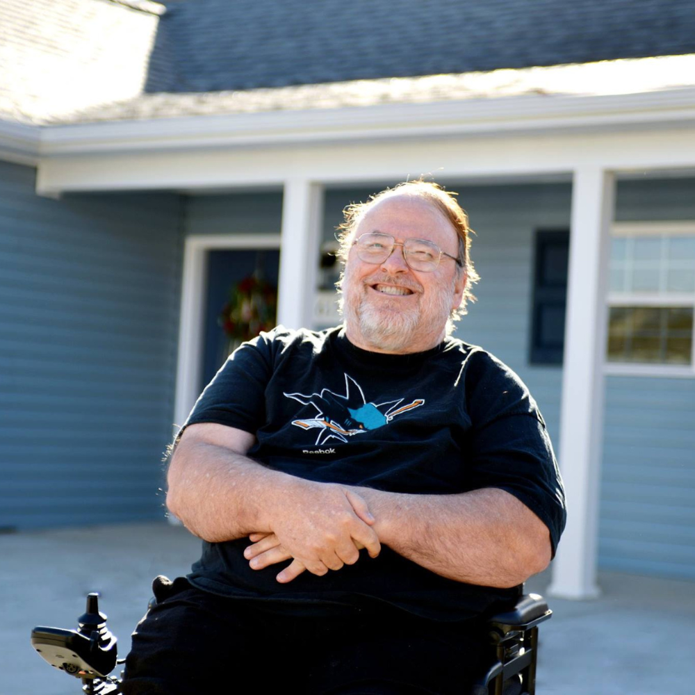 A middle-aged caucasian man is smiling and sitting in a wheelchair outside of a blue single-story.