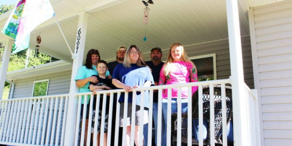 A family of six is standing outside on the front porch of a house.