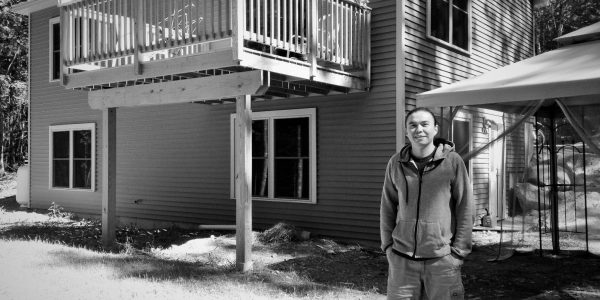 A young adult man is standing outside of a two-story home.