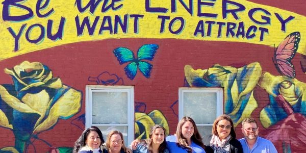 A group of six people are standing in front of a painted mural that says "Be the energy you want to attract". There are roses and butterflies on the mural.