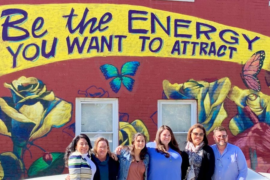 A group of six people are standing in front of a painted mural that says "Be the energy you want to attract". There are roses and butterflies on the mural.