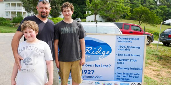 A caucasian father and two young sons are standing and smiling outside in a neighborhood.