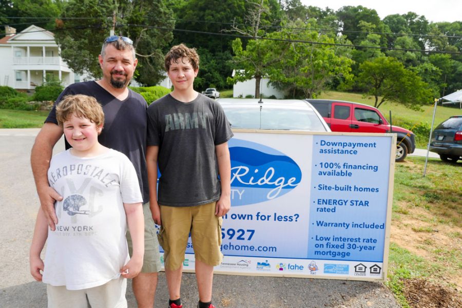A caucasian father and two young sons are standing and smiling outside in a neighborhood.