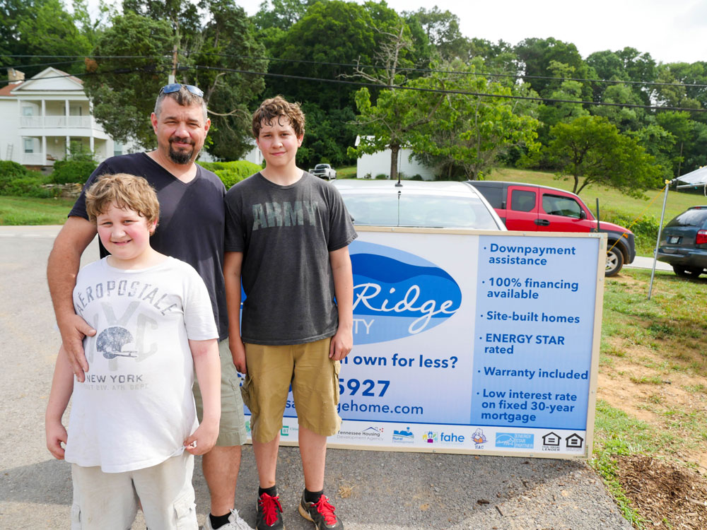 A caucasian father and two young sons are standing and smiling outside in a neighborhood.