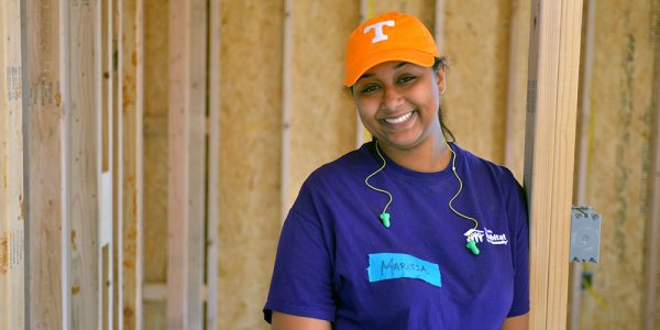 A woman is smiling and standing in a doorframe of an unfinished home. She is wearing a Habitat for Humanity t-shirt.