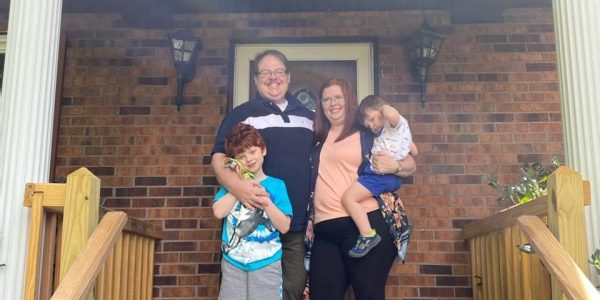 A family of four, including two parents and two young children, are standing on a front porch outside of their home.