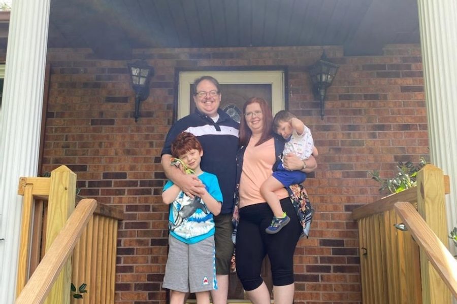 A family of four, including two parents and two young children, are standing on a front porch outside of their home.