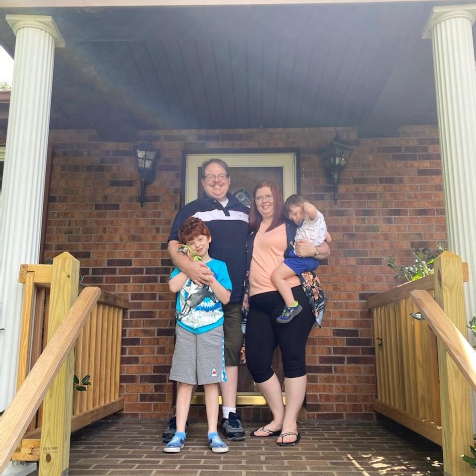 A family of four, including two parents and two young children, are standing on a front porch outside of their home.