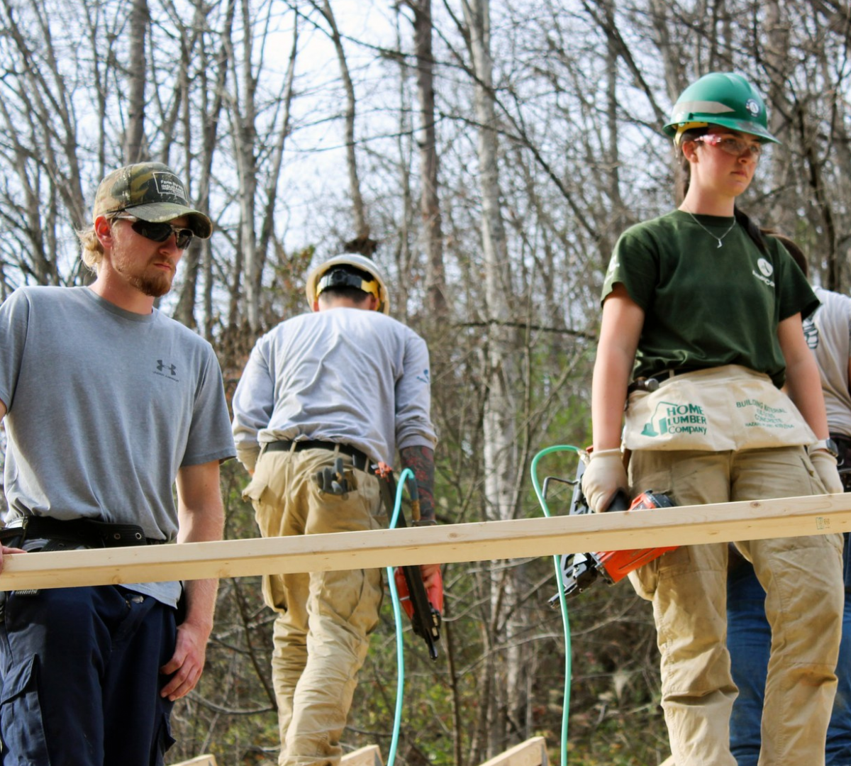 Two caucasian people are standing holding lumber and construction tools outside. There are trees and other construction works behind them.