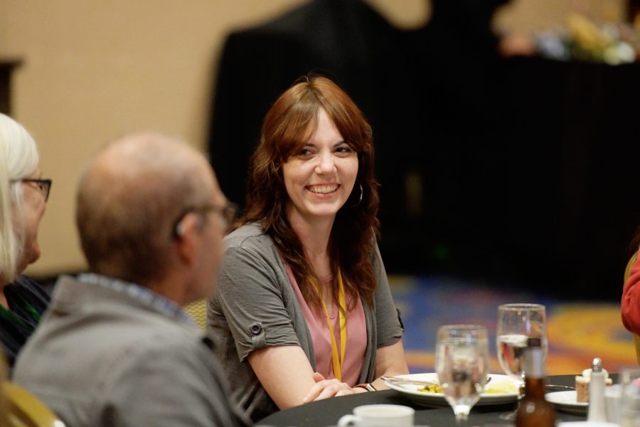 A caucasian woman is seated at a table smiling looking at someone else seated at the table. They are inside a ballroom at an annual event.