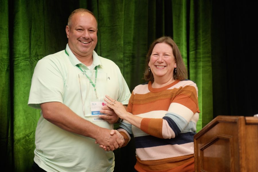 Two caucasian Fahe members are standing on a stage smiling and shaking hands.
