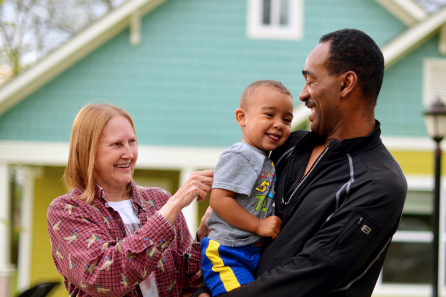 An older African American man is smiling holding a young African American boy in his arms. There is an older caucasian woman standing beside them smiling. Behind them is a blue and yellow home.