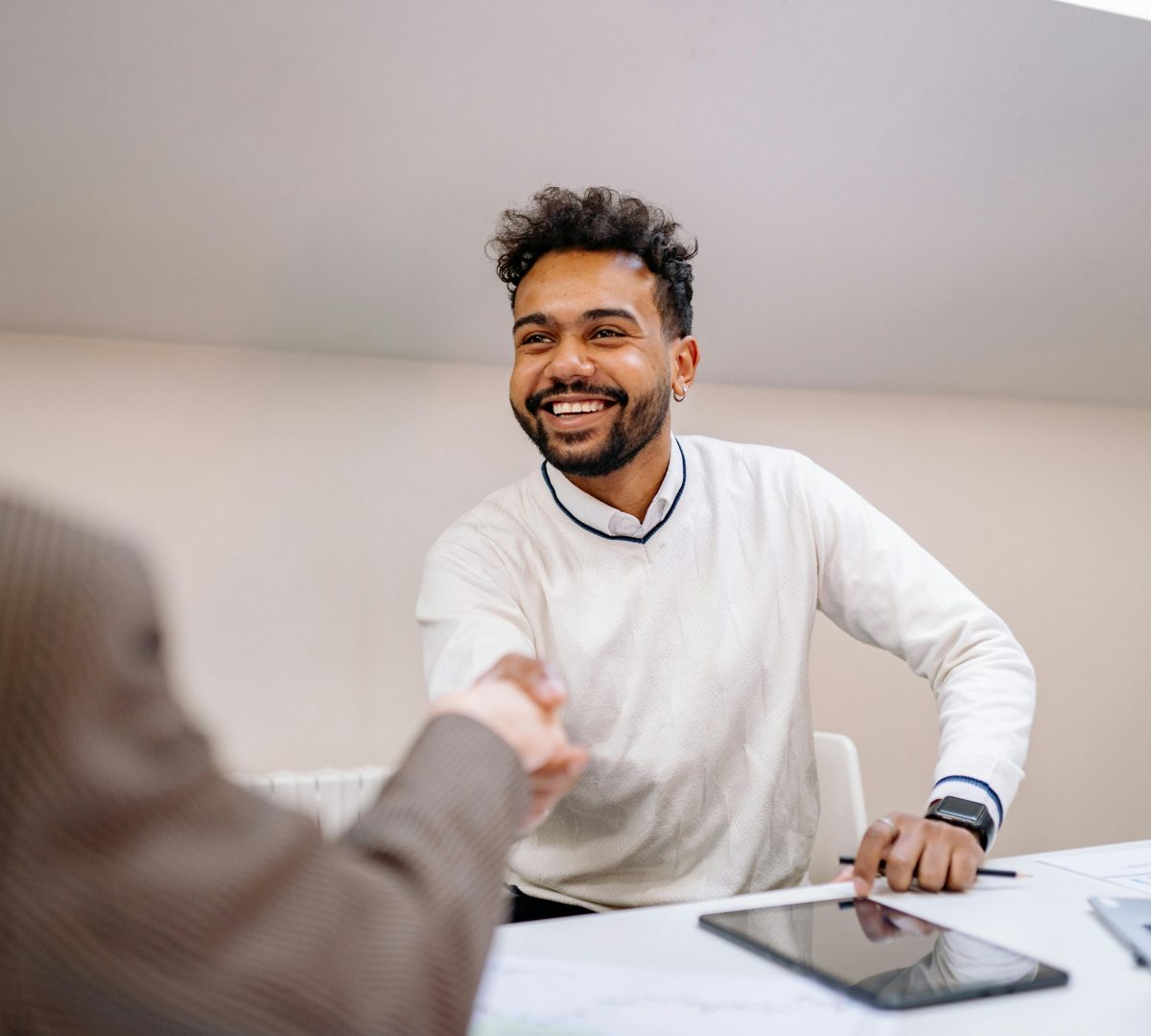A young adult man of color smiling and is shaking hands with a professional in an office setting.