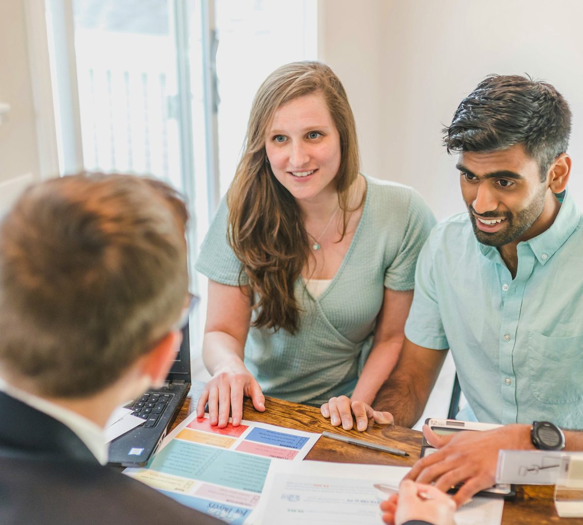 A young couple is smiling sitting across from a lending professional at a desk. They have their hands on the table with paper and pens beside them.