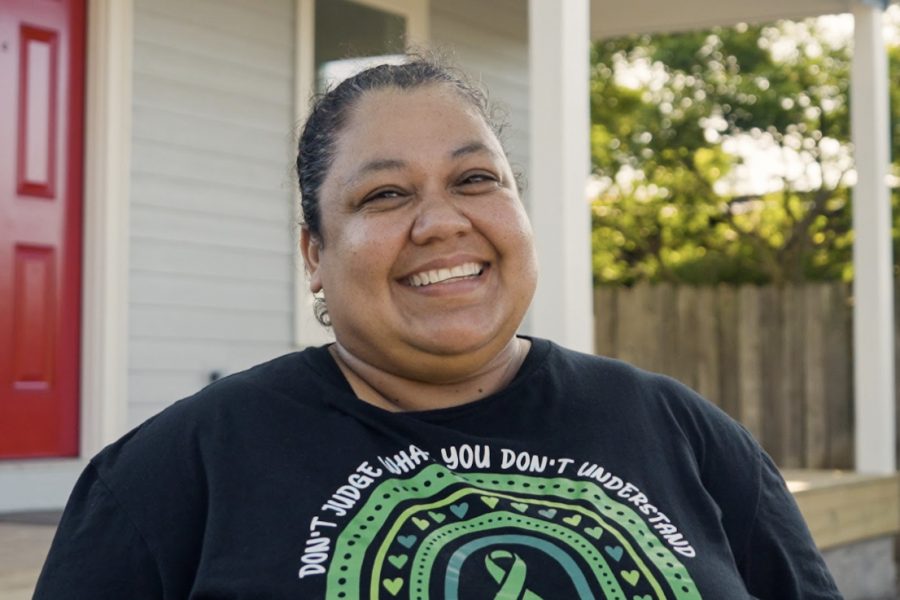 A woman of color is smiling standing outside of a house. The house is gray with a red door.