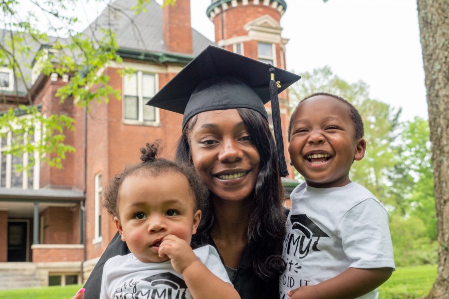 A young African American mother is smiling holding her two smiling children outside. She is wearing a black university cap and gown.