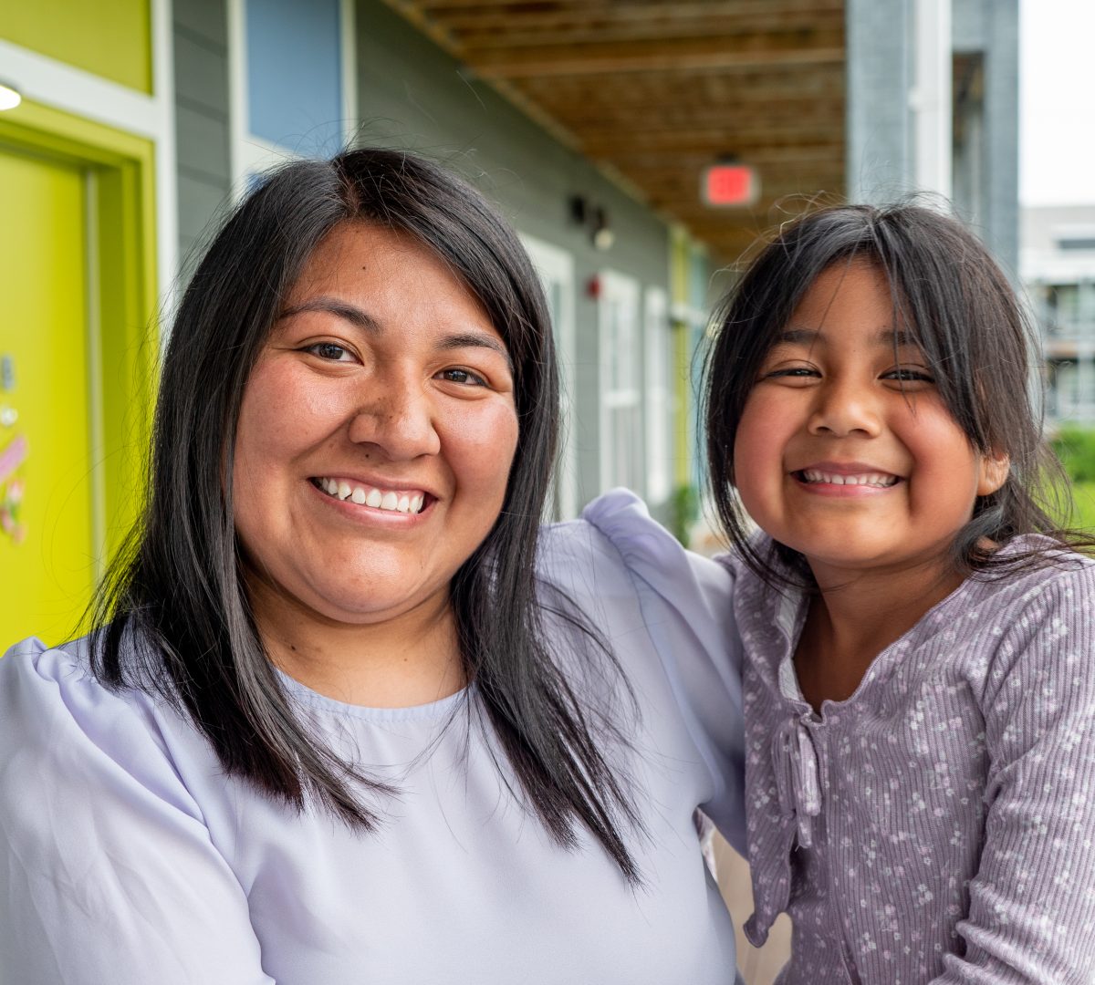 A woman of color is smiling holding her daughter. They are standing outside of a mutli-story apartment complex.