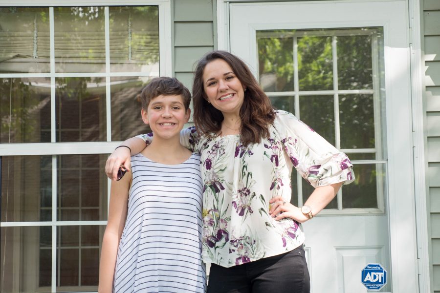A caucasian woman is smiling and standing outside of a home with her arm around her teenage child.