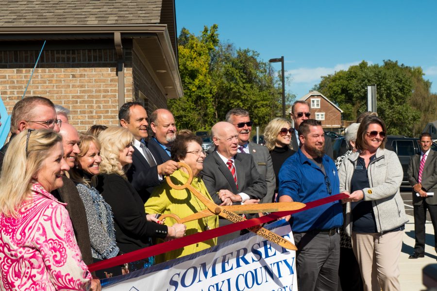A group of people are standing outside smiling at a ribbon cutting event. Two people are holding a red ribbon and a two people are holding a giant pair of wooden scissors open about to cut the ribbon.
