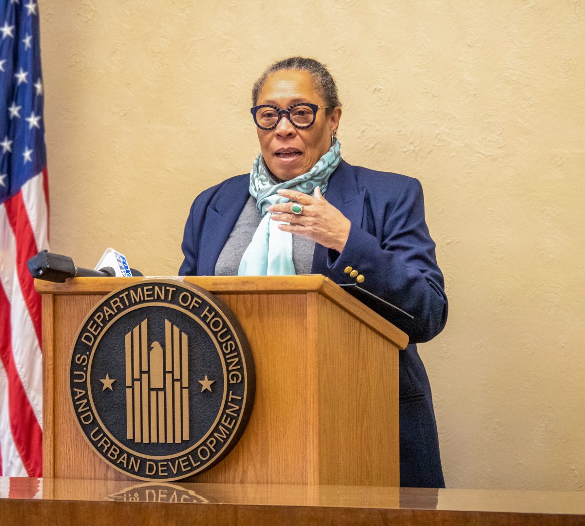 An African American woman is speaking at a podium that has U.S Department of Housing and Urban Development written on the front it. There is a tan wall behind her and an American flag.